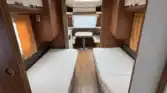 Interiér obytného vozu prodej karavanu se dvěma samostatnými lůžky po obou stranách, dřevěnými skříňkami a malou jídelnou se stolem a oknem na vzdáleném konci. Podlaha má pruhovaný vzor připomínající dřevo.