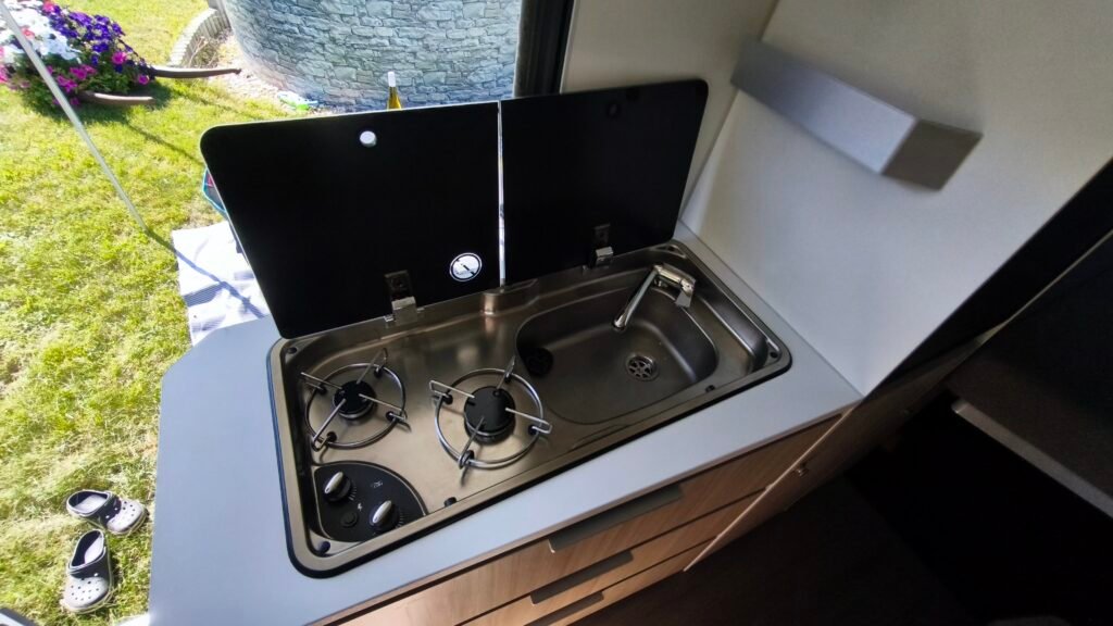 Obrázek zobrazuje kuchyňský kout v obytném autě s dřezem a dvouplotýnkovým vařičem.
