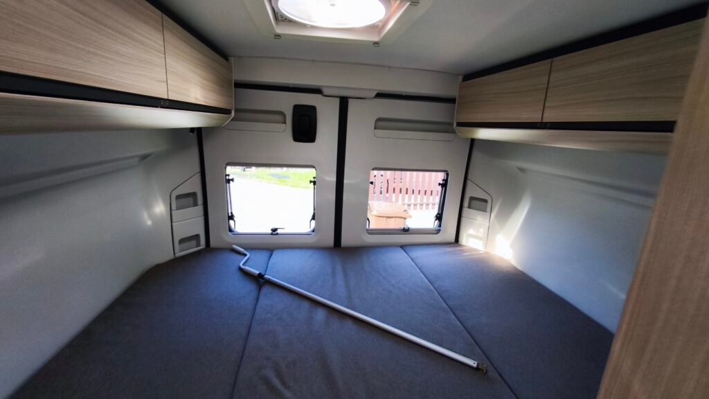 Obrázek interiéru karavanu, světlé dřevěné skříňky a prostorná postel pro pohodlné spaní.