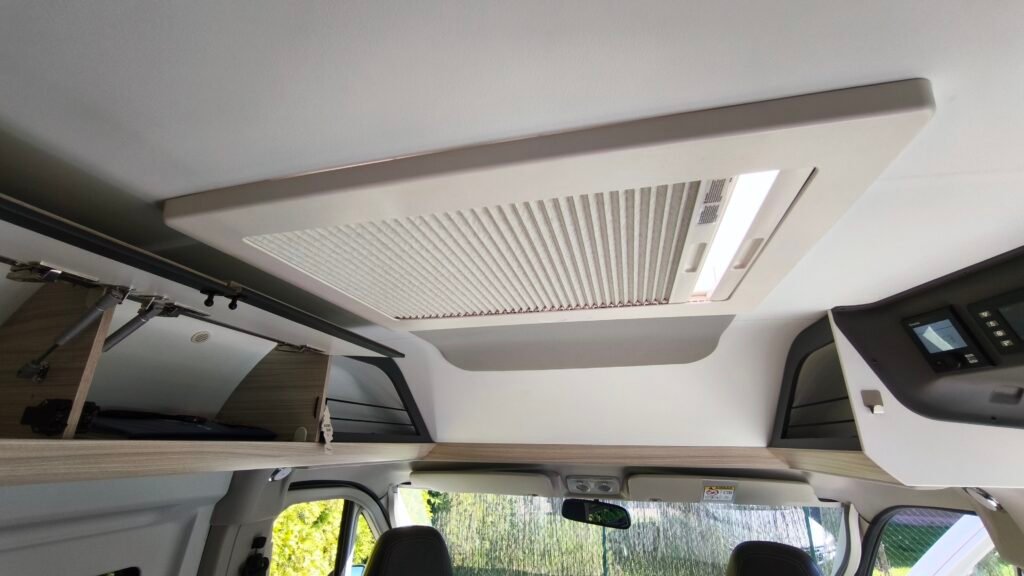 Obrázek interiéru obytného auta, včetně klimatizace a úložných prostor. Elegantní design a funkčnost pro pohodlné cestování.