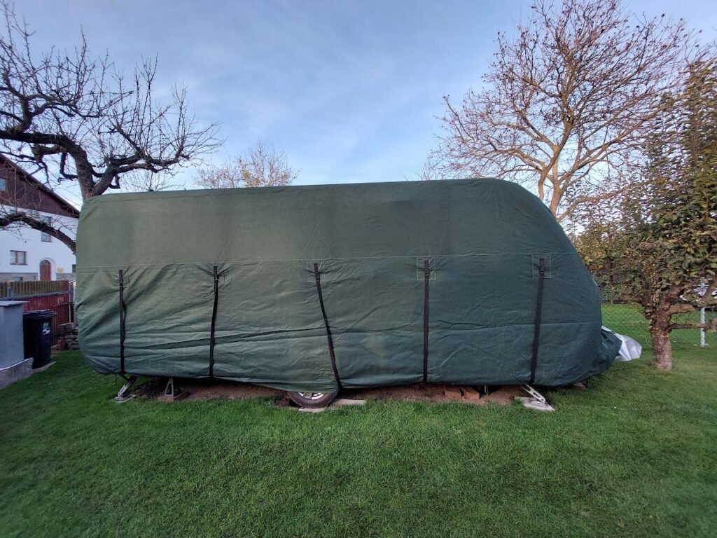 Na obrázku je zakrytý karavan, uložený na trávě. Není vidět značka ani model.