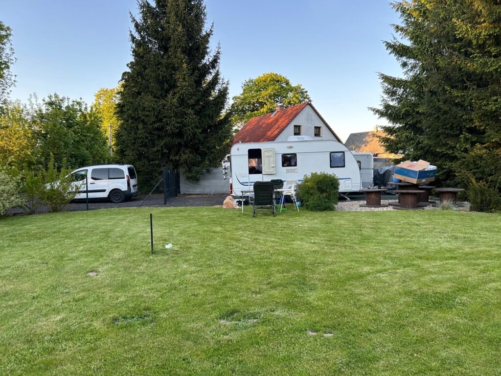 Na obrázku je bílý karavan v zahradě s posezením a okolní zelení.