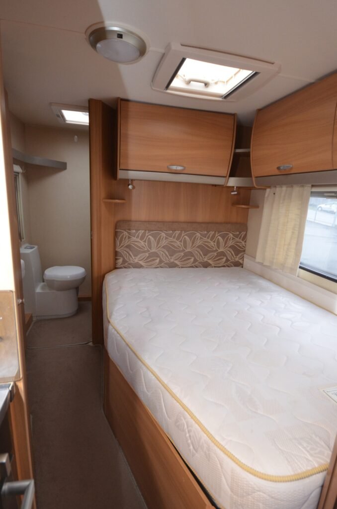 Interiér karavanu s manželskou postelí a toaletou. Moderní dřevěná výbava. Ideální pro pohodlné cestování.