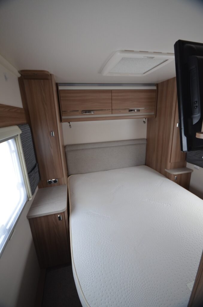 Obrázek interiéru karavanu s komfortní postelí a úložným prostorem v dřevěném stylu.