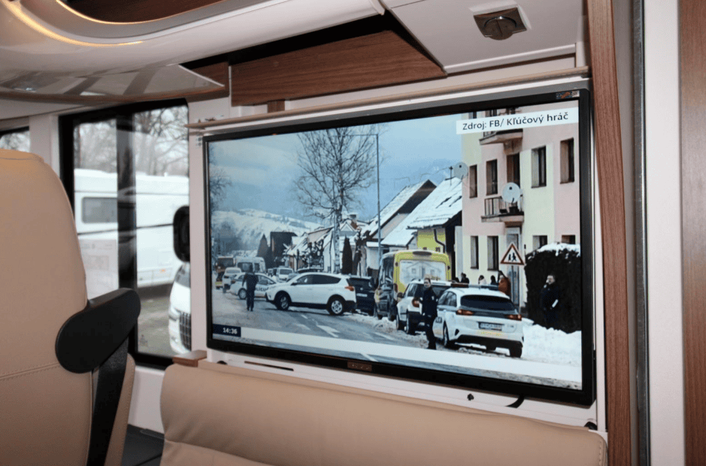 Na obrázku je obytné auto s televizí, nachází se v interiéru. V pozadí jsou vidět další vozidla.