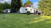 Na obrázku je karavan zaparkovaný na zahradě, vedle obytňáku. Vzadu je stříšková konstrukce a stůl s lavičkami.