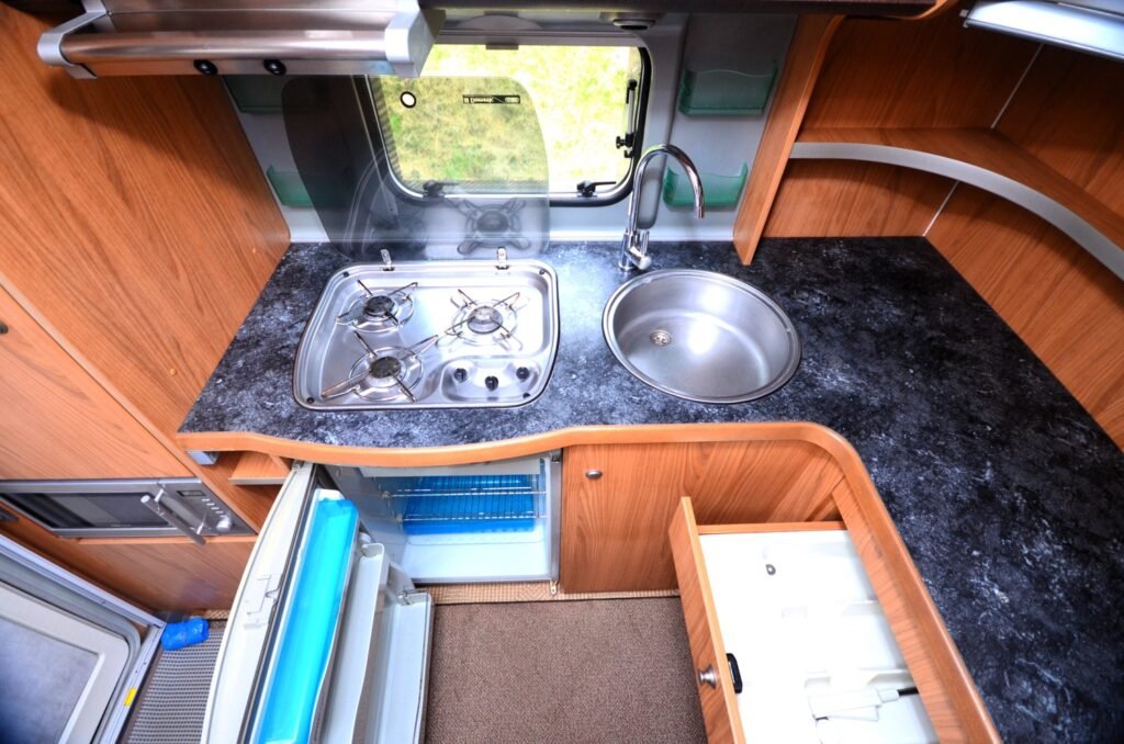 Obrázek zobrazuje kuchyňský kout v karavanu, vybavený sporákem, dřezem a ledničkou.