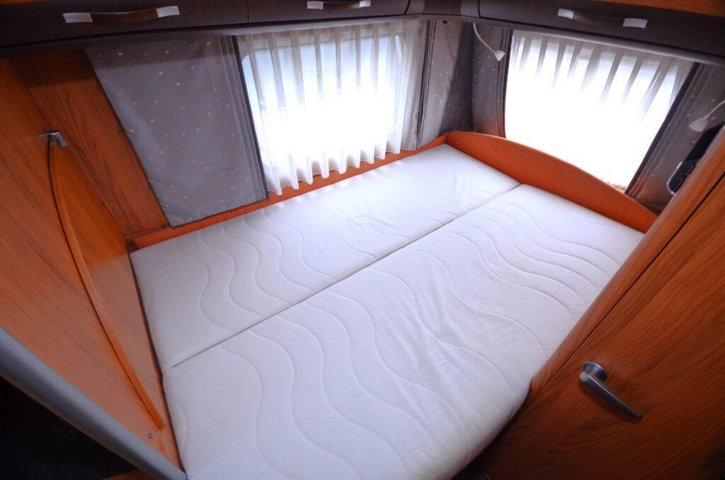 Interiér karavanu s pohodlnou postelí a dřevěnými prvky, ideální pro relaxaci na cestách.