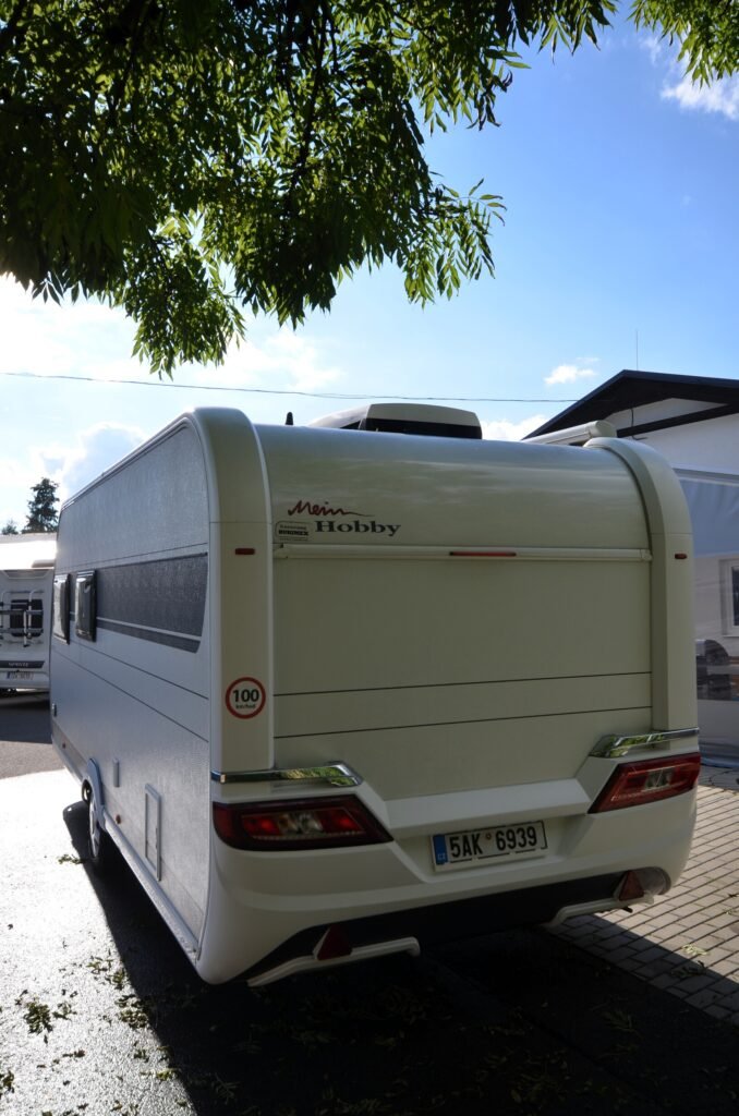 Karavan Hobby, bílý design, parkovaný pod stromy. Ideální pro pohodlné cestování a kempování.