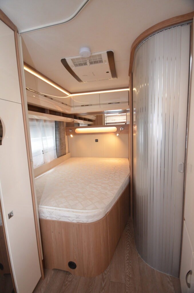 Interiér moderního obytného auta s manželskou postelí a úložným prostorem. Elegantní design a pohodlné osvětlení.