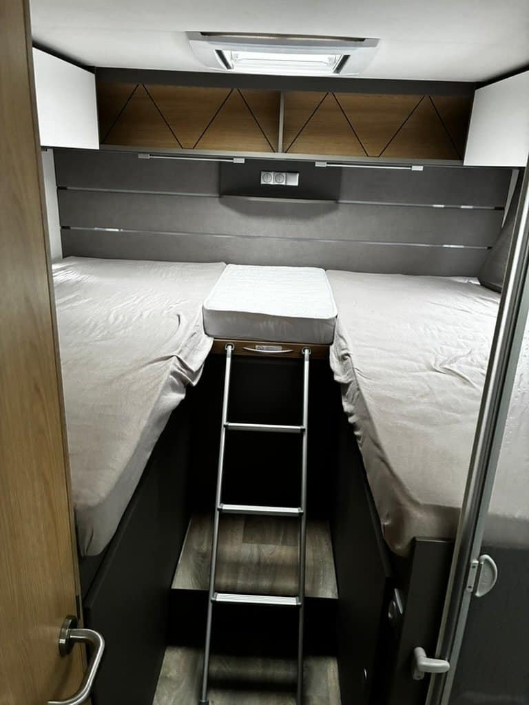Interiér obytného auta s dvoulůžkem, schůdky a úložným prostorem. Moderní design a pohodlí na cestách.
