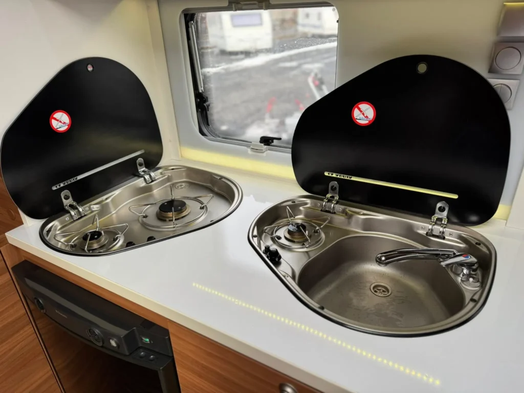 Obrázek ukazuje kuchyňský kout v obytném autě s dvojumývadlem a vařičem. Značka a model nejsou vidět.