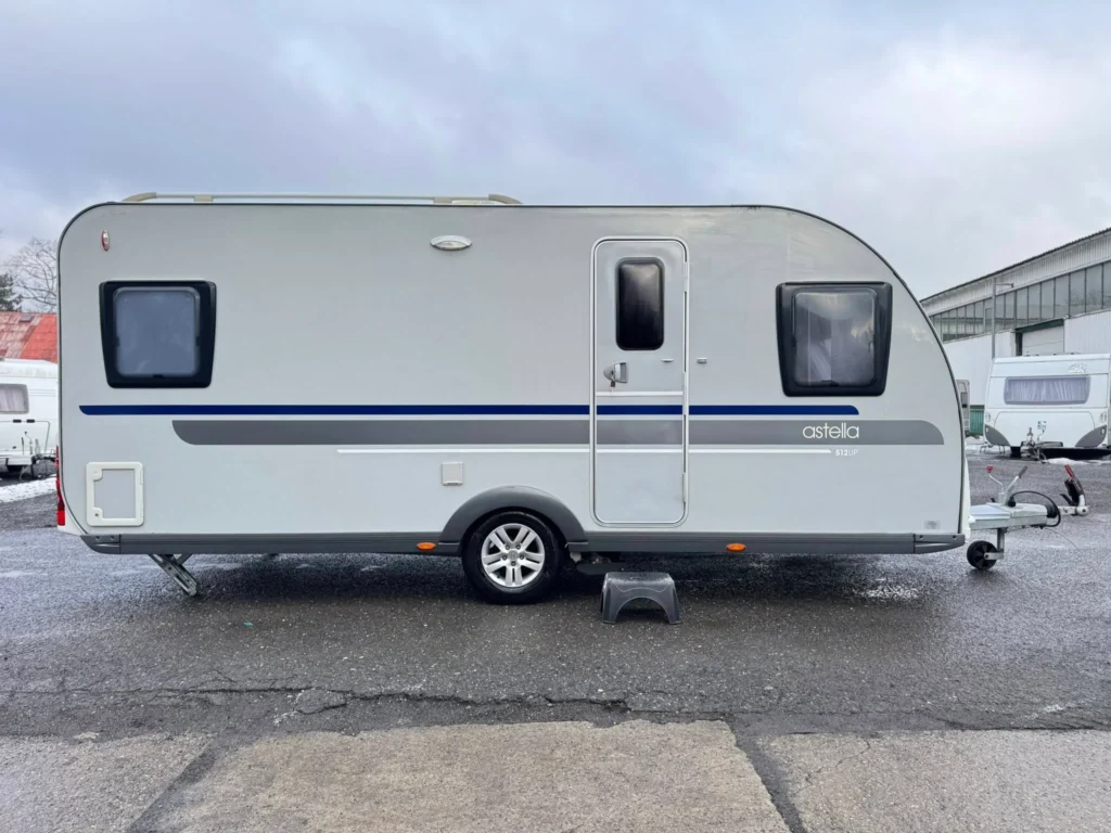 Na zpevněné ploše stojí moderní stříbrnošedý karavan s jednou nápravou. U jeho vstupních dveří je malý černý schůdek a v pozadí je vidět další karavan připravený k prodeji nebo k prohlídce interiéru.