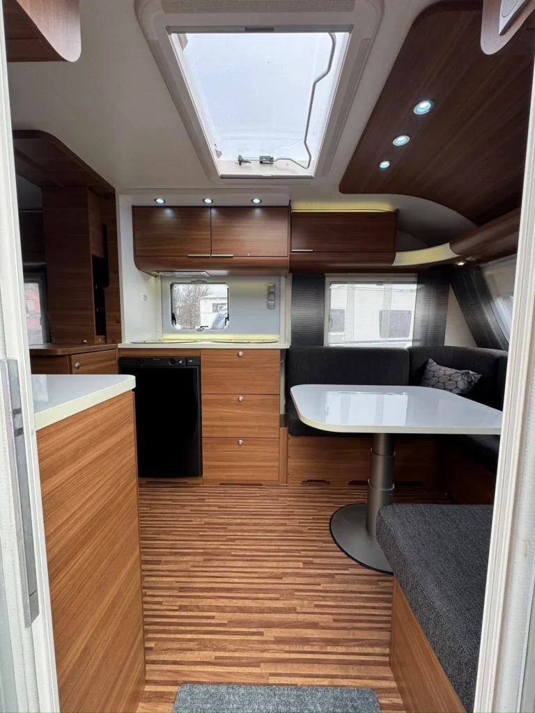 Obrázek interiéru karavanu s dřevěnými prvky a moderním vybavením. Relaxační prostor se stolkem a kuchyňkou.