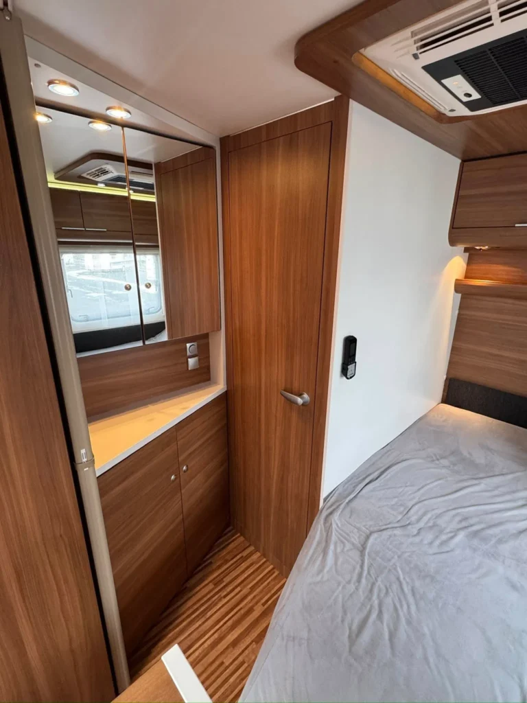 Interiér obytného přívěsu s dřevěnými prvky, zrcadlem a manželskou postelí. Viditelné skříně a osvětlení.