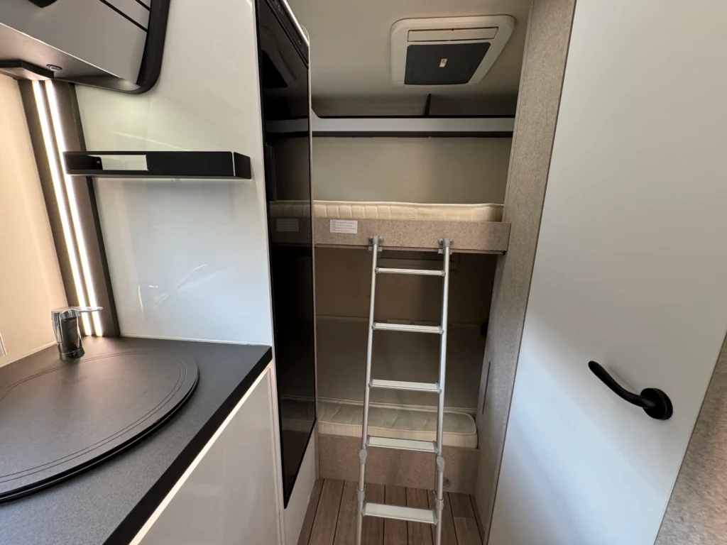 Obrázek interiéru obytného auta s kuchyňským koutem a dvoupodlažním spaním. Komfortní a moderní design.