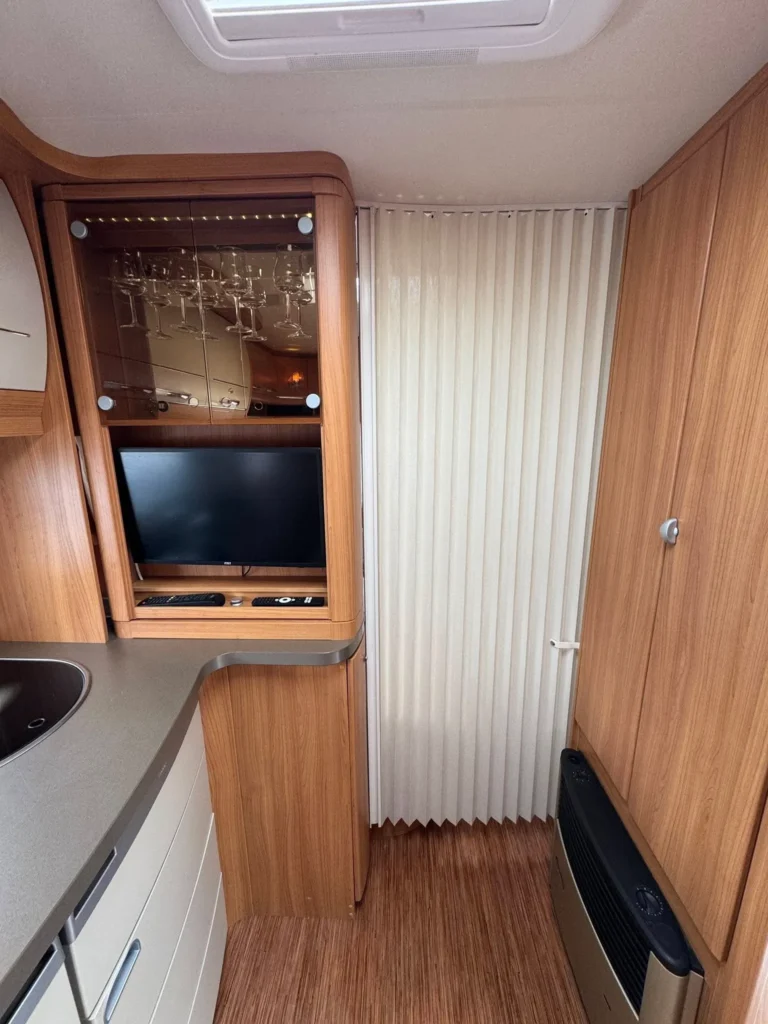 Interiér moderního obytného auta s dřevěným nábytkem, TV a úložným prostorem pro sklenice. Komfortní a útulný design.