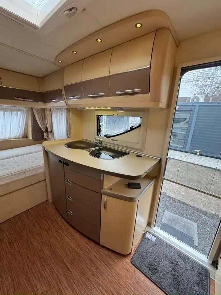 Interiér obytného přívěsu s kuchyňskou linkou, skříňkami a prostorem pro spaní. Vkusné barevné provedení.