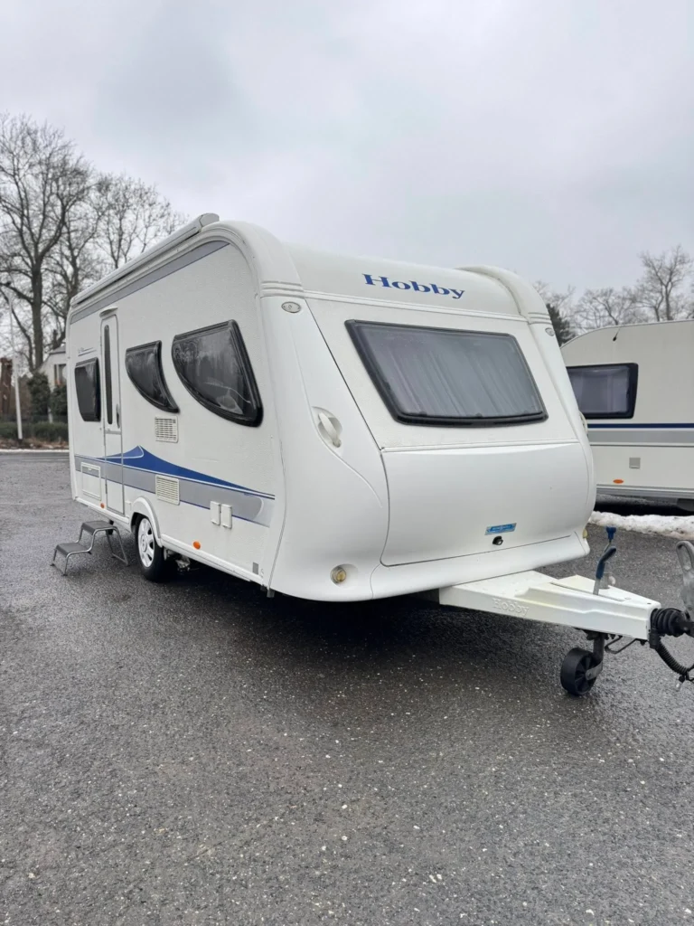 Karavan značky Hobby, bílý s modrými pruhy, parkovaný na šedém asfaltu.