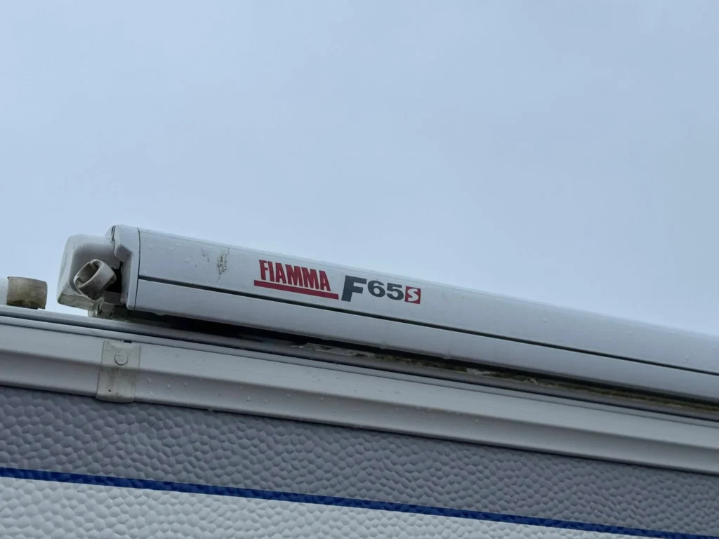 Obrázek zobrazuje markýzu Fiamma F65S namontovanou na karavanu nebo obytném přívěsu.