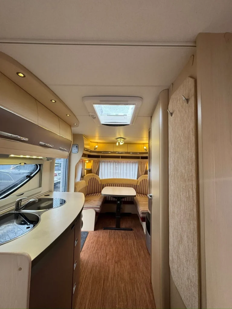 Interiér obytného přívěsu s kuchyňským koutem a jídelním prostorem, příjemná atmosféra s dřevěnou podlahou a oknem.