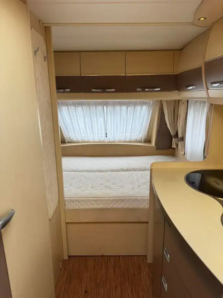 Interiér obytného přívěsu s dřevěnou podlahou a kuchyňským koutem, viditelné lůžko a okna se záclonami.