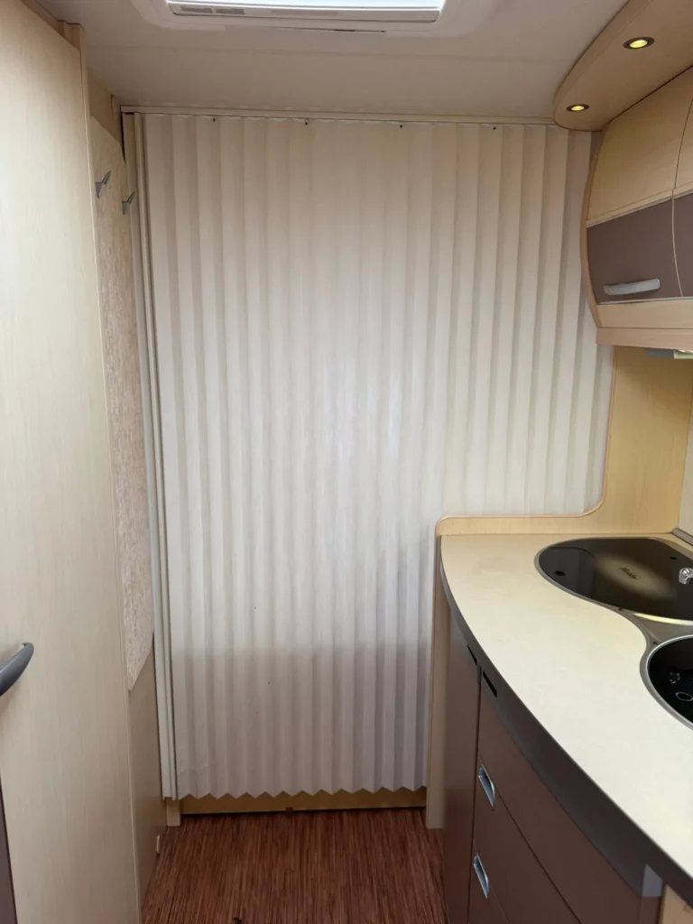 Interiér karavanu s kuchyňským koutem, skříněmi a žaluziemi. Moderní design v neutrálních barvách.