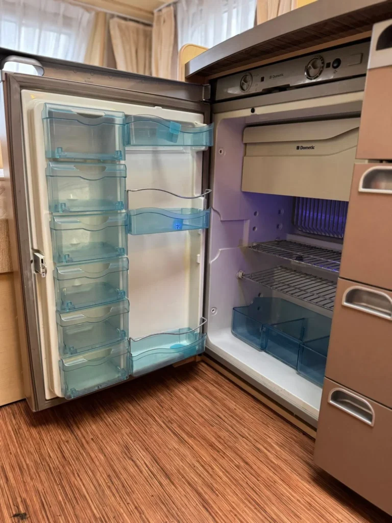 Na obrázku je interiér karavanu s lednicí Dometic, viditelné modré zásobníky a police.