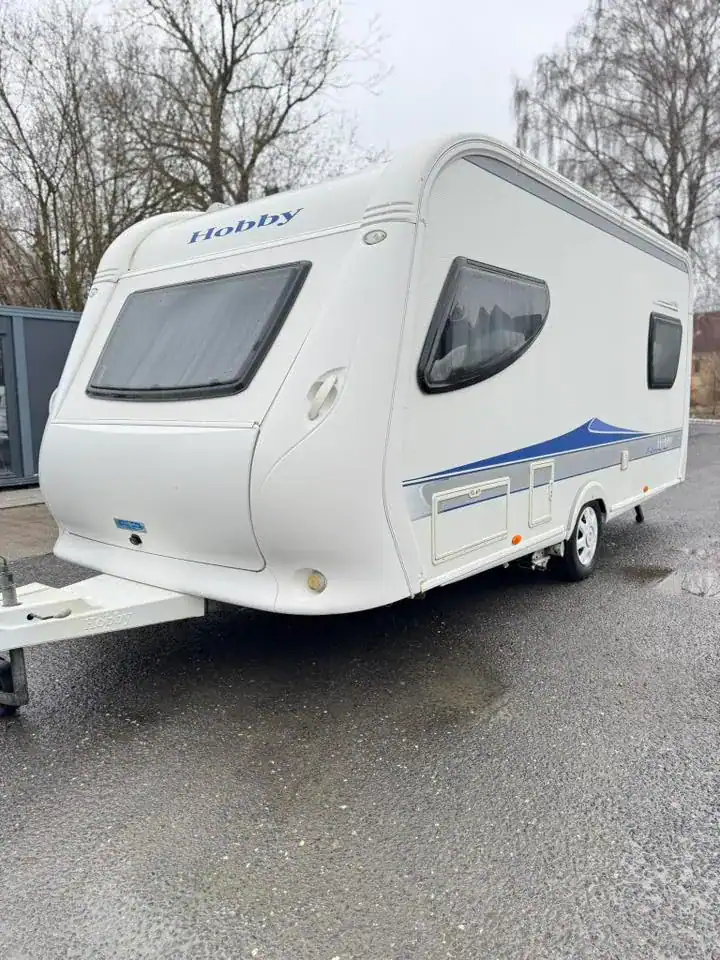 Na obrázku je obytný přívěs značky Hobby, bílá karoserie s modrými detaily.