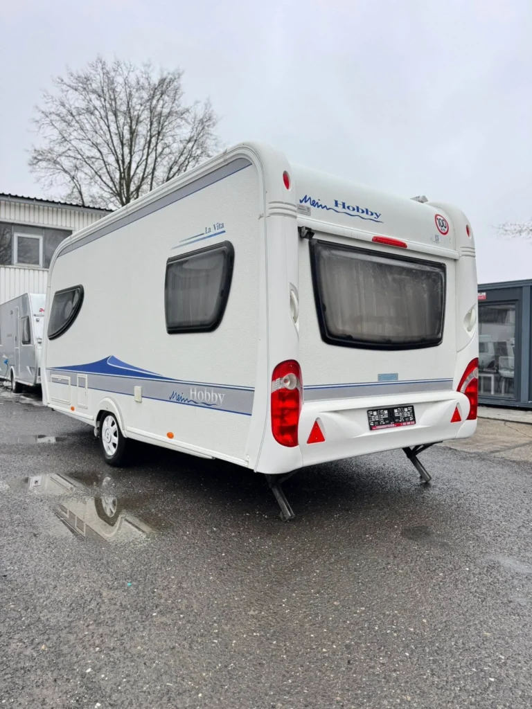 Obrázek zobrazuje obytný přívěs značky Hobby, model La Vita, zaparkovaný na mokré zemi.