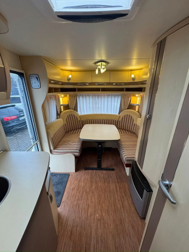 Interiér karavanu s kuchyňským koutem, sedací soupravou a stolem. Osvětlení je příjemné a prostorné uspořádání.