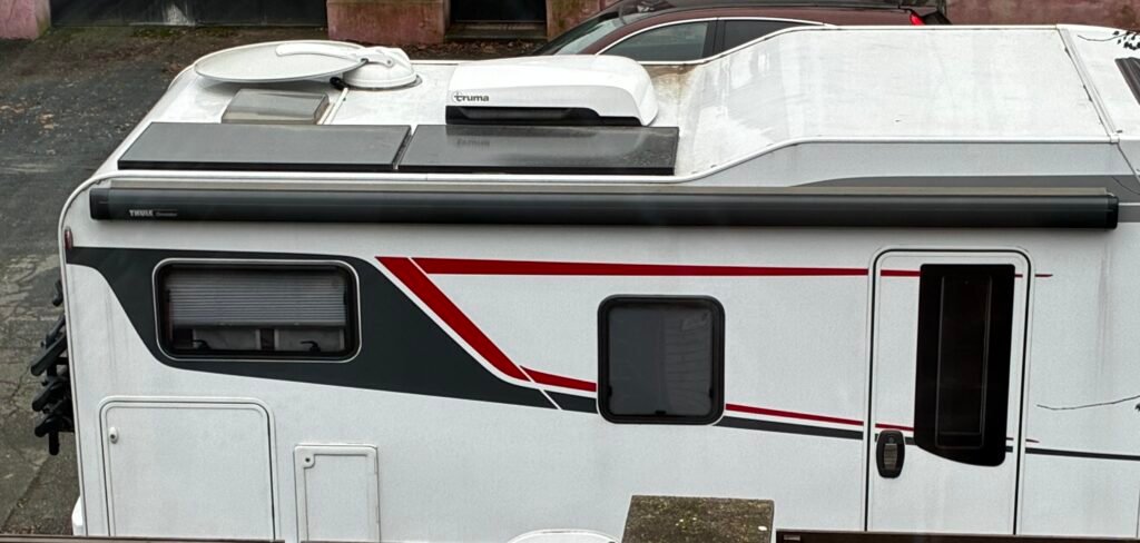 Na obrázku je karavan s bílým exteriérem, červenými a černými pruhy. Viditelný je střešní ventilátor a markýza Thule.