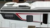 Na obrázku je karavan s bílým exteriérem, červenými a černými pruhy. Viditelný je střešní ventilátor a markýza Thule.