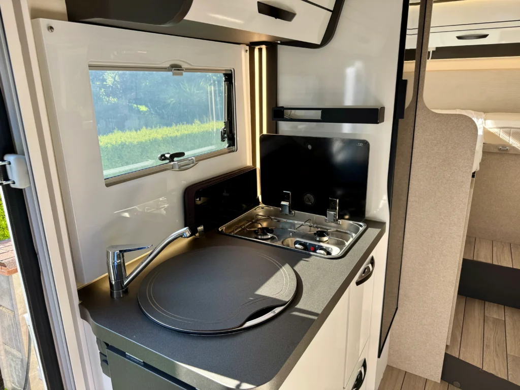 Interiér obytného auta s moderním kuchyňským koutem, varnou deskou a dřezem. Prostorné a světlé prostředí.