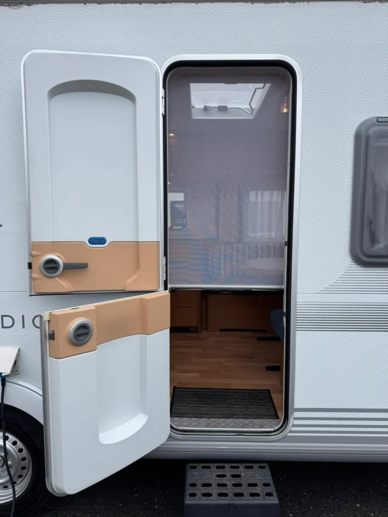 Obrázek ukazuje karavan s otevřenými dveřmi, připravený na dobrodružství. Vybavení uvnitř je dobře osvětlené.
