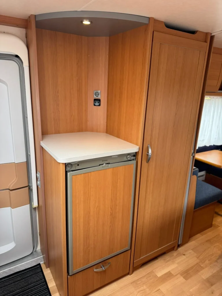 Interiér karavanu s lednicí a úložným prostorem. Dřevěné prvky dodávají teplý vzhled a útulnost.