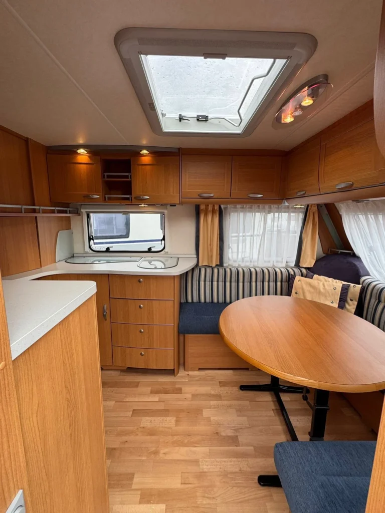 Interiér karavanu s dřevěnými skříňkami a sedačkami. Komfortní stůl a kuchyňský kout pro příjemné pobyty.