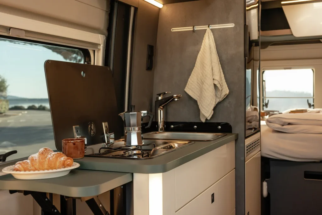 Obrázek interiéru obytného auta s kuchyňským koutem, nádobím a pohodlným spaním. Ideální pro kempování a relaxaci.