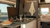 Obrázek interiéru obytného auta s kuchyňským koutem, nádobím a pohodlným spaním. Ideální pro kempování a relaxaci.