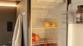 Obrázek zobrazuje interiér obytného vozu s chladničkou plnou ovoce a nápojů.