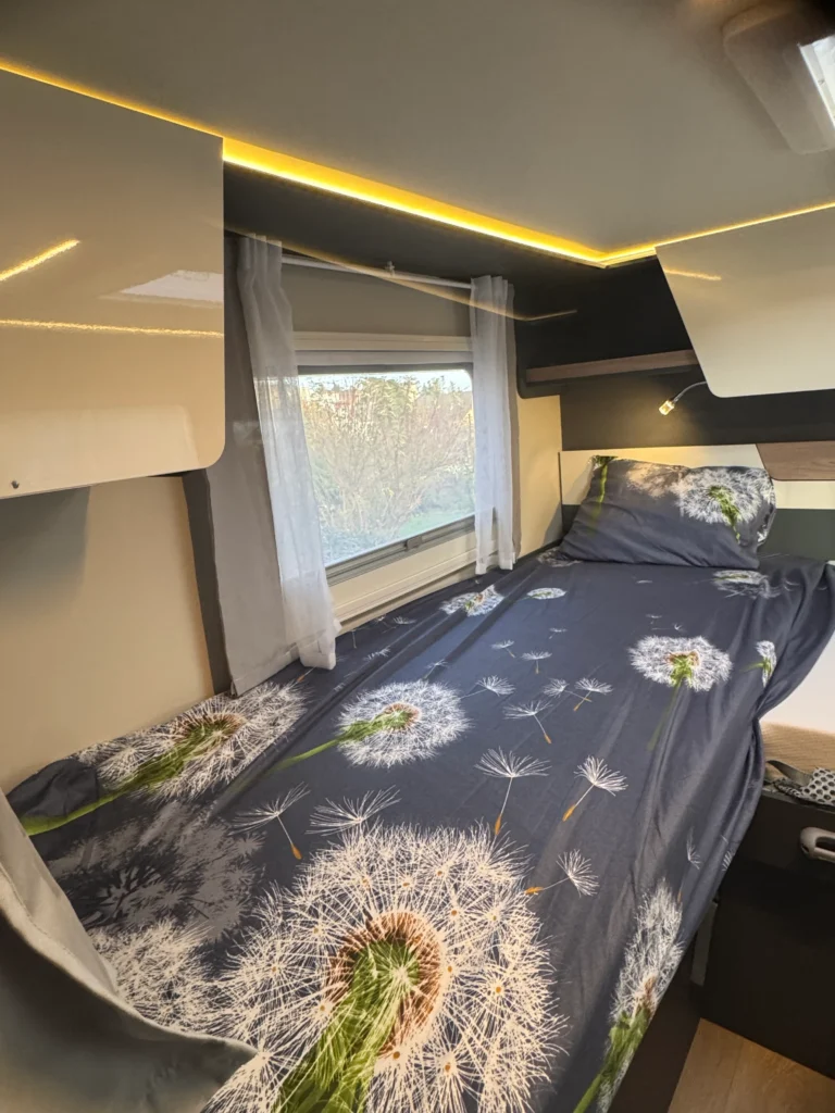 Obrázek představuje interiér obytného vozu se stylovým ložním prádlem a útulným osvětlením.