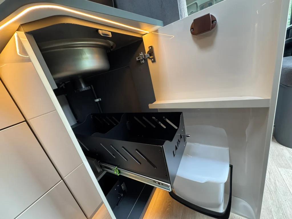 Obrázek zobrazuje část interiéru karavanu, konkrétně úložný prostor s kovovým dřezem a přihrádkou na odpadky.
