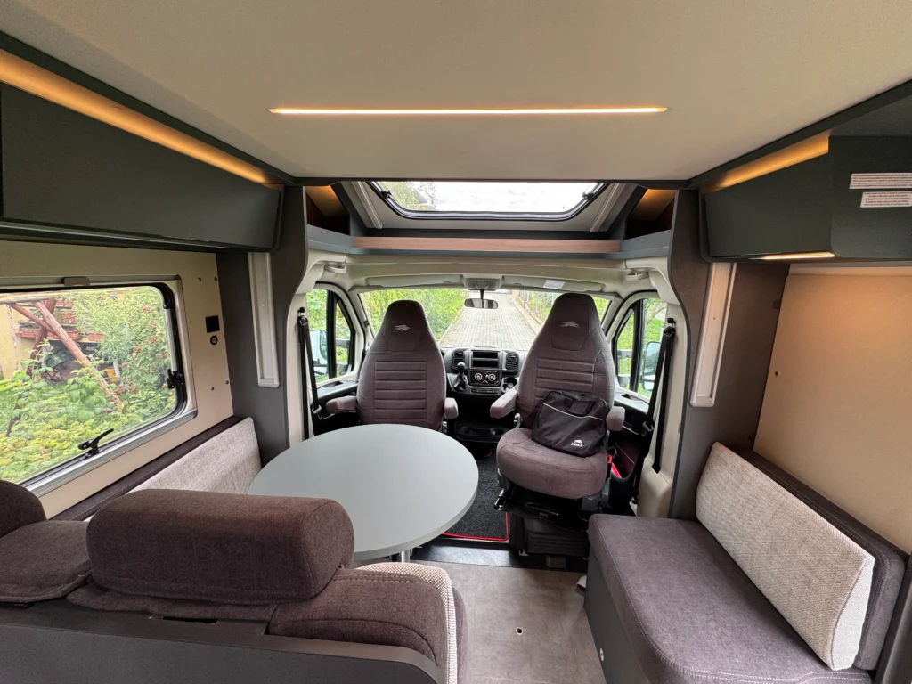 Interiér karavanu s prostorem pro sezení a jídelním stolem. Viditelné pohodlné sedačky a moderní osvětlení.