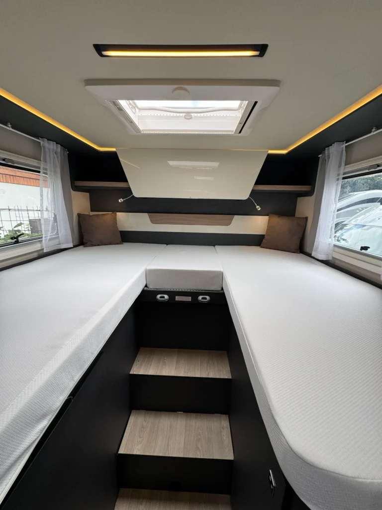 Interiér obytného přívěsu, s rozloženou postelí a moderním osvětlením. Detailní pohled na prostor a schody.