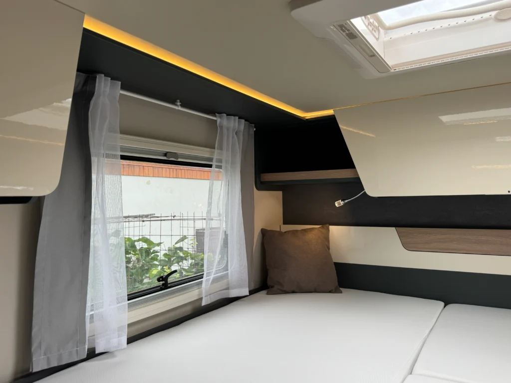 Interiér moderního obytného přívěsu s manželskou postelí, oknem a ambientním osvětlením.