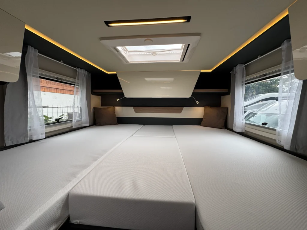 Interiér obytného přívěsu, přehledné uspořádání s bílými matracemi, okna s bílými závěsy a moderním osvětlením.