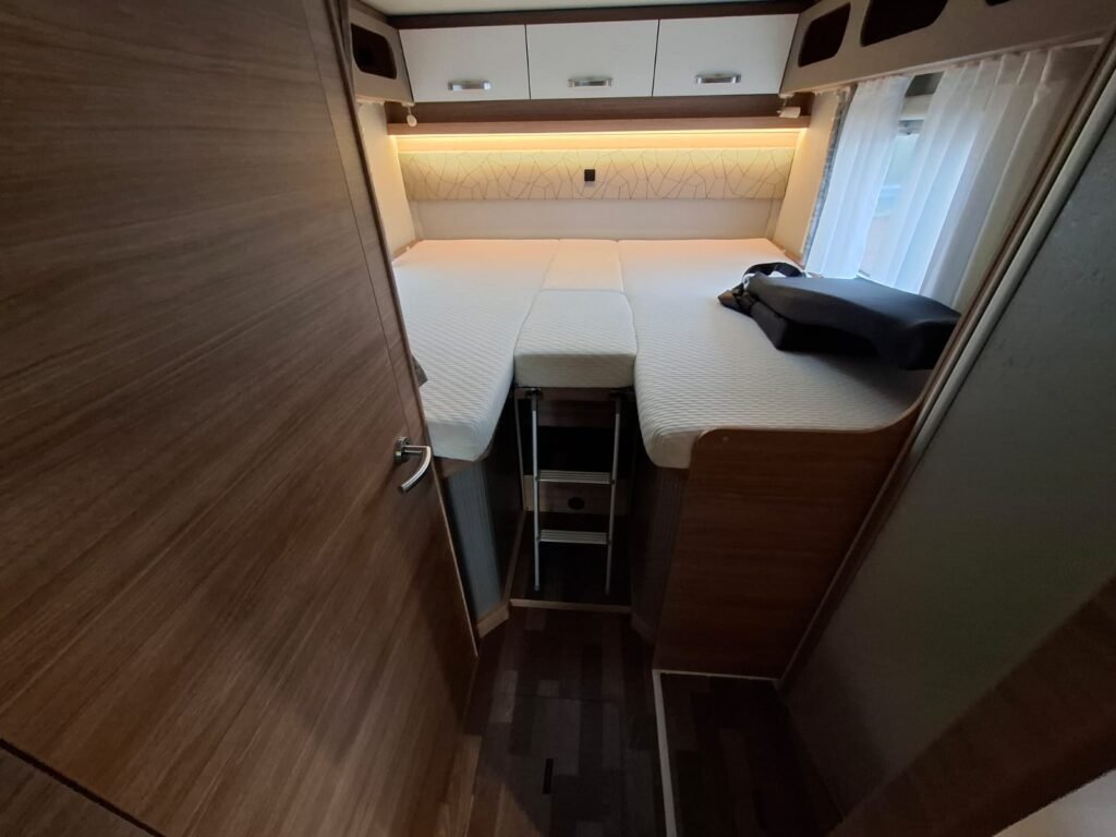 Interiér obytného auta s lůžkem a úložným prostorem, moderní design s dřevěnými prvky.