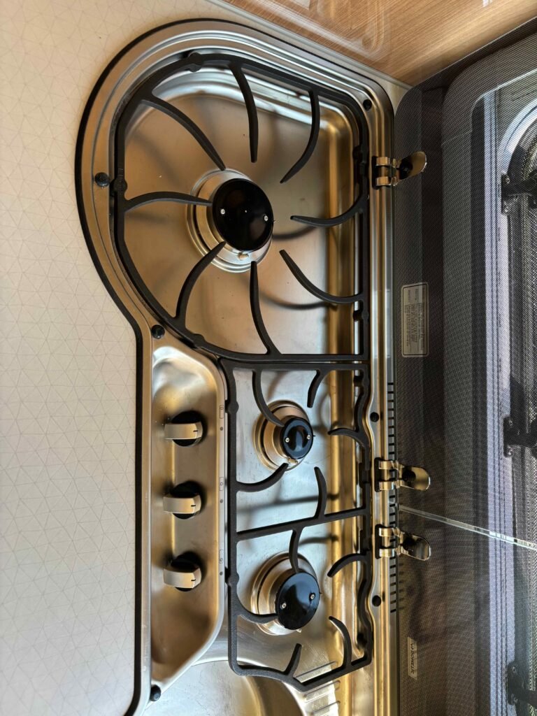 Na obrázku je varná deska z obytného přívěsu alebo karavanu. Zobrazuje čtyři hořáky a ovládací knoflíky.