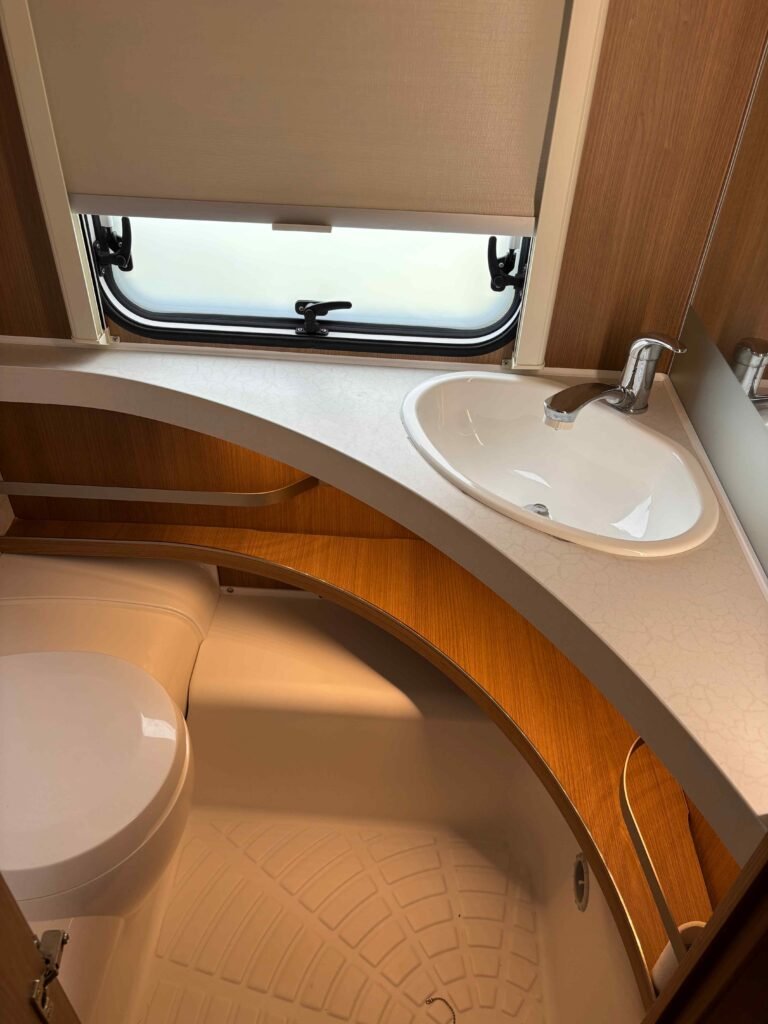 Interiér karavanu nebo obytného přívěsu s umyvadlem a toalettou, dřevěné obložení, okno s roletou.