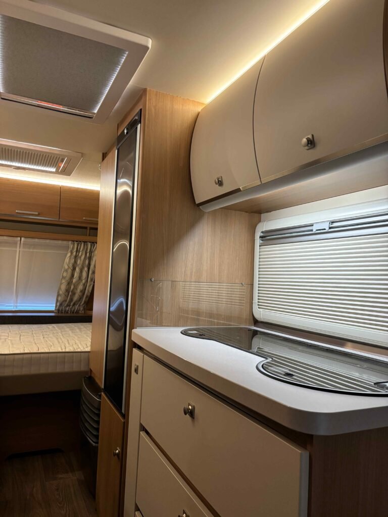 Interiér karavanu s kuchyňkou, lednicí a manželskou postelí; moderní design s dřevěnými prvky a dobrým osvětlením.